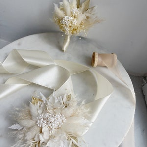 May include: Two white dried flower bouquets with pampas grass and white flowers. The bouquets are on a white marble surface with a white satin ribbon.