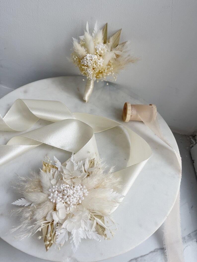 May include: Two white dried flower bouquets with pampas grass and white flowers. The bouquets are on a white marble surface with a white satin ribbon.