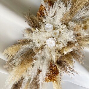 May include: A dried floral arrangement with white pampas grass, brown pampas grass, and white roses. The arrangement is accented with dried hydrangeas in a beige color.