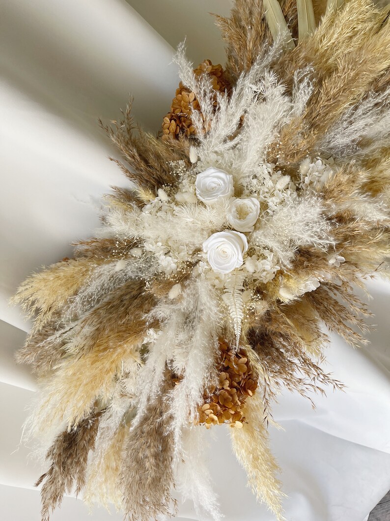 May include: A dried floral arrangement with white pampas grass, brown pampas grass, and white roses. The arrangement is accented with dried hydrangeas in a beige color.