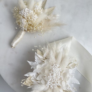 May include: Two white dried flower arrangements with pampas grass and white baby's breath. The arrangements are on a white marble surface.