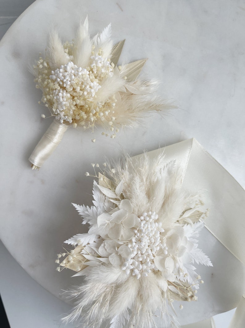 May include: Two white dried flower arrangements with pampas grass and white baby's breath. The arrangements are on a white marble surface.