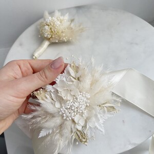 May include: A white and cream dried flower bouquet with a white satin ribbon. The bouquet is made with pampas grass, dried hydrangeas, and other dried flowers.