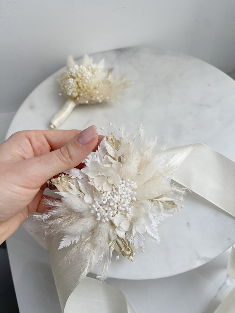May include: A white and cream dried flower bouquet with a white satin ribbon. The bouquet is made with pampas grass, dried hydrangeas, and other dried flowers.