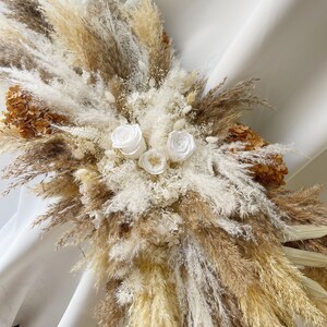 May include: A dried floral arrangement featuring white pampas grass, beige pampas grass, and white roses. The arrangement is in a vase and is on a white background.