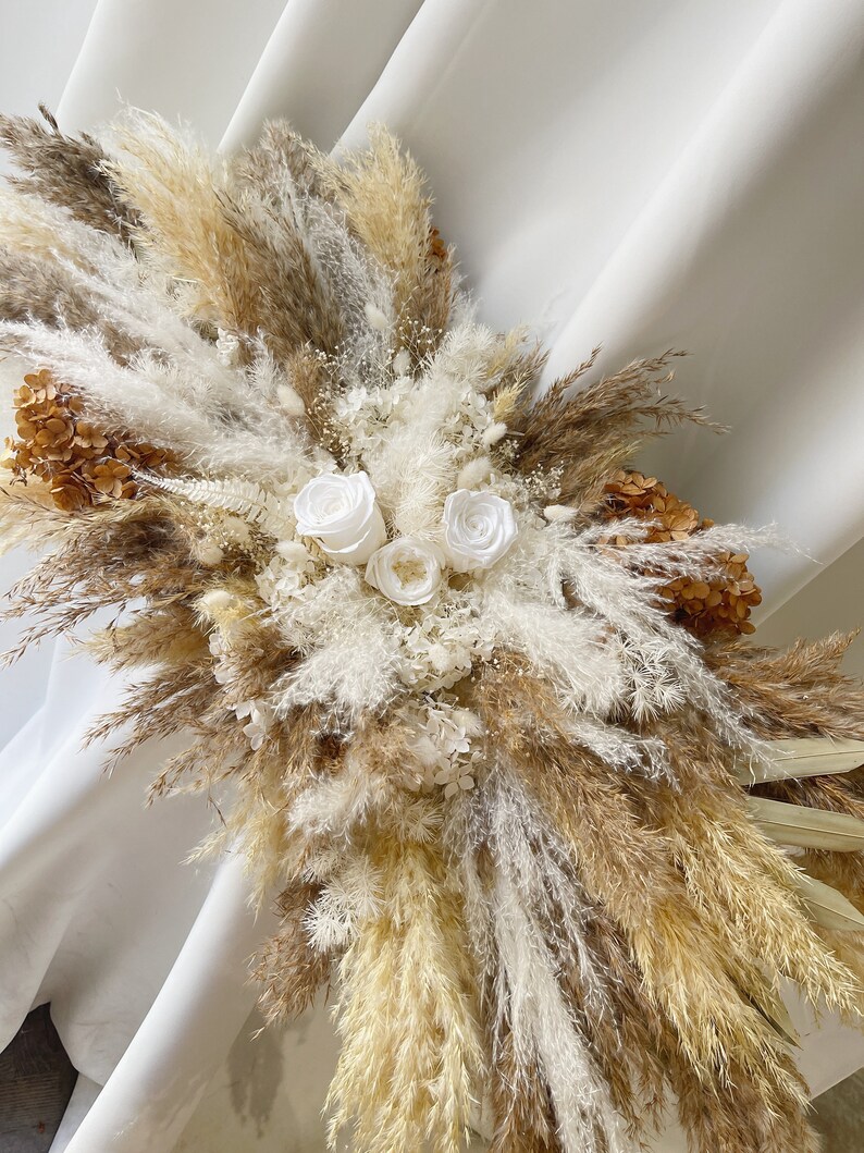 May include: A dried floral arrangement featuring white pampas grass, beige pampas grass, and white roses. The arrangement is in a vase and is on a white background.