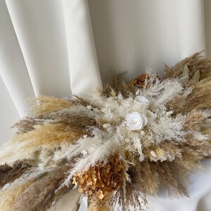 May include: A dried floral arrangement with white and brown pampas grass, white roses, and brown hydrangeas.