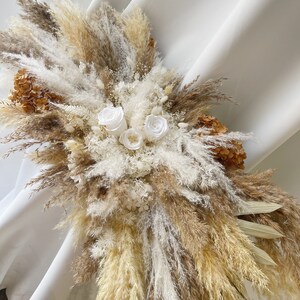 May include: A dried floral arrangement featuring a mix of pampas grass, dried flowers, and white roses. The arrangement is in shades of beige, brown, and white.