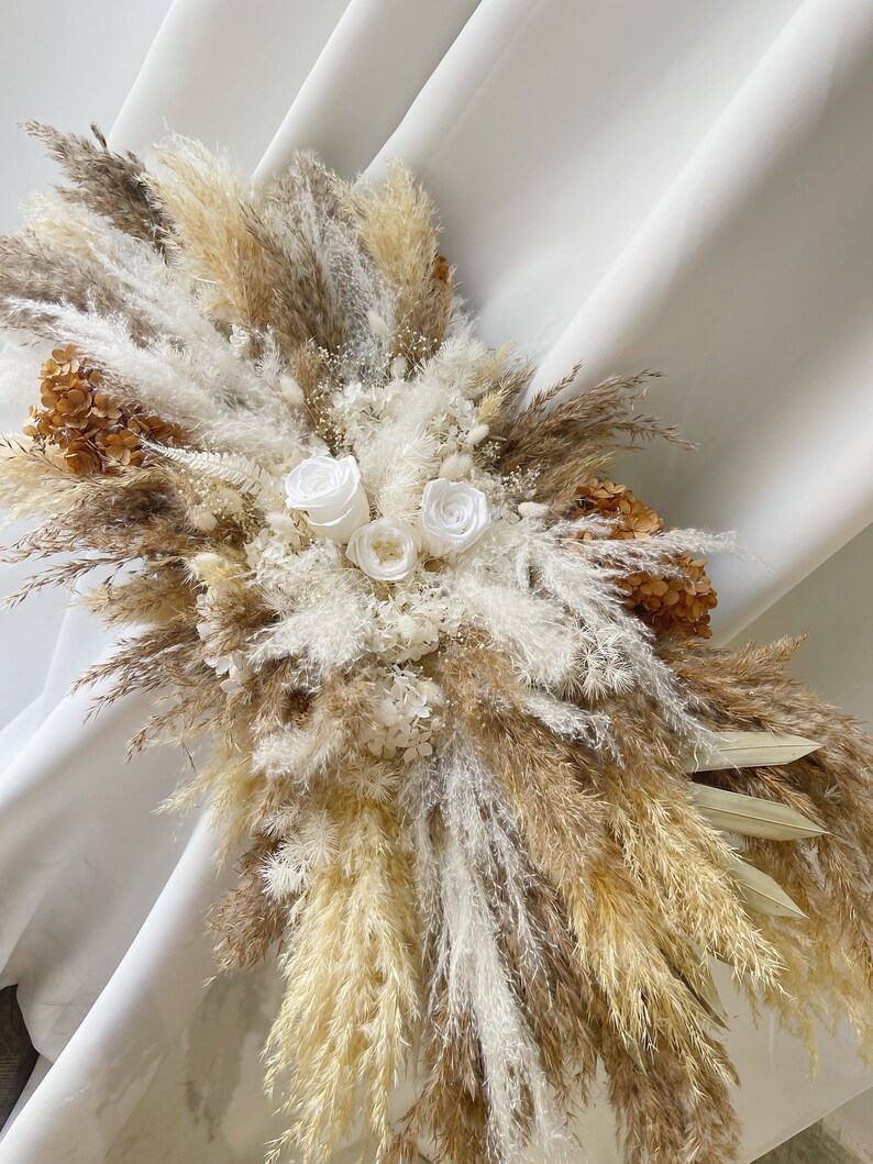 May include: A dried floral arrangement featuring a mix of pampas grass, dried flowers, and white roses. The arrangement is in shades of beige, brown, and white.