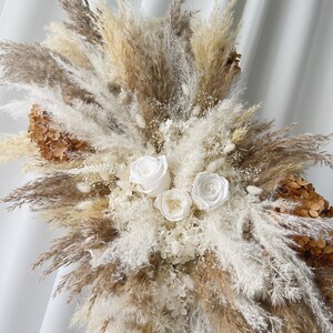 May include: A dried floral arrangement featuring white roses, pampas grass, and other dried flowers in shades of beige and white.