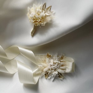 May include: Two white and gold dried flower arrangements. One is a small bouquet with a white ribbon, the other is a larger arrangement with a white satin ribbon.