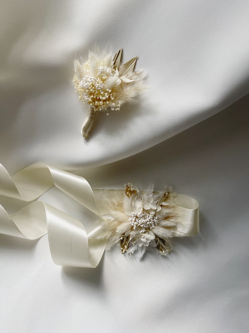 May include: Two white and gold dried flower arrangements. One is a small bouquet with a white ribbon, the other is a larger arrangement with a white satin ribbon.