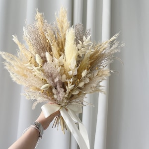 May include: A bouquet of dried pampas grass, bunny tails, and other dried flowers in shades of beige, cream, and brown, tied with a white satin ribbon.