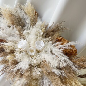 May include: A bouquet of dried pampas grass, white dried flowers, and white roses. The bouquet is arranged in a fan shape and is against a white background.