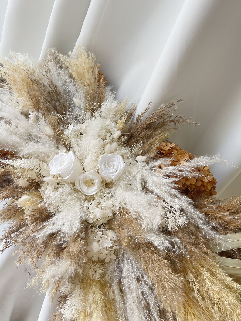 May include: A bouquet of dried pampas grass, white dried flowers, and white roses. The bouquet is arranged in a fan shape and is against a white background.