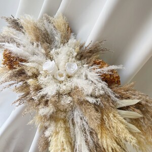 May include: A dried floral arrangement with white and beige pampas grass, white roses, and dried flowers. The arrangement is displayed against a white backdrop.
