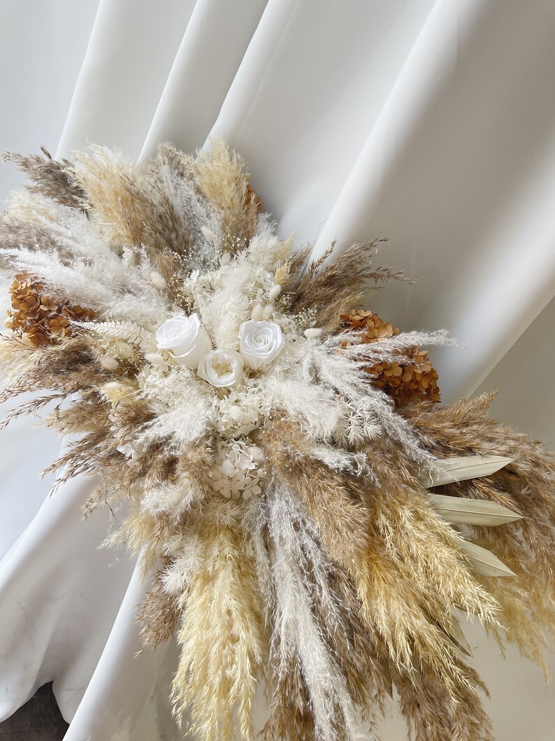 May include: A dried floral arrangement with white and beige pampas grass, white roses, and dried flowers. The arrangement is displayed against a white backdrop.