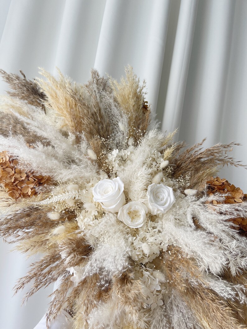May include: A dried floral arrangement featuring white roses, white pampas grass, and brown pampas grass.