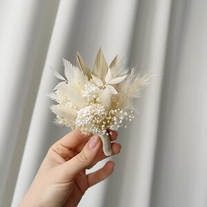 May include: A small bouquet of dried flowers in shades of white and beige, tied with a white ribbon. The bouquet includes pampas grass, bunny tails, and other dried flowers.