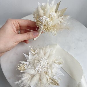 May include: Two white dried flower bouquets with pampas grass and white baby's breath. The bouquets are tied with white ribbon.