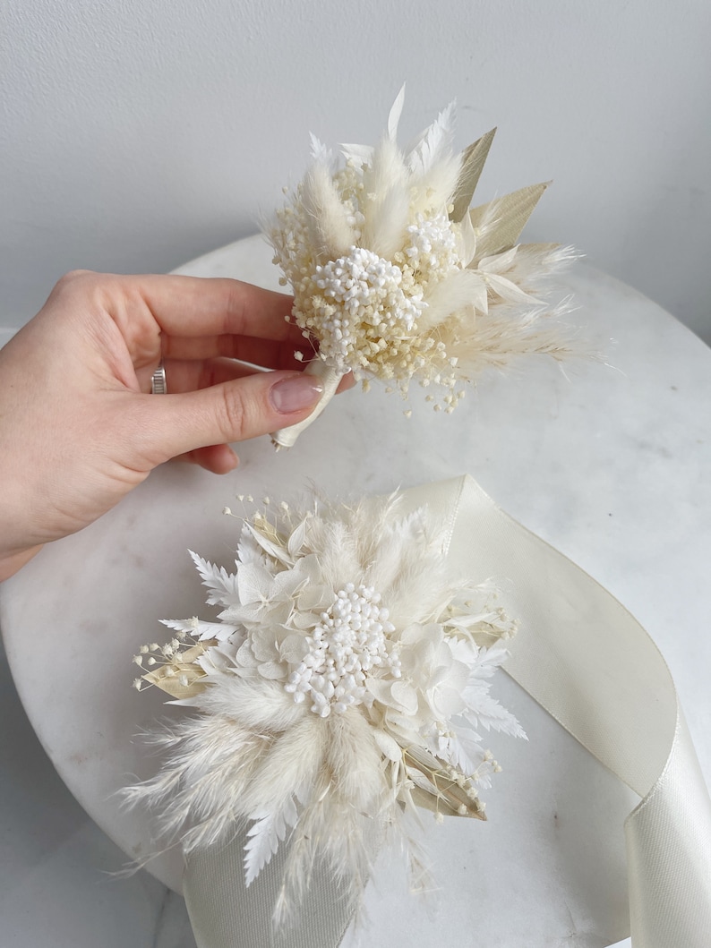 May include: Two white dried flower bouquets with pampas grass and white baby's breath. The bouquets are tied with white ribbon.