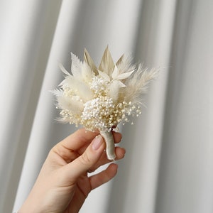 May include: A small bouquet of dried flowers in shades of white and beige, tied with a white ribbon. The bouquet includes pampas grass, baby's breath, and other dried foliage.