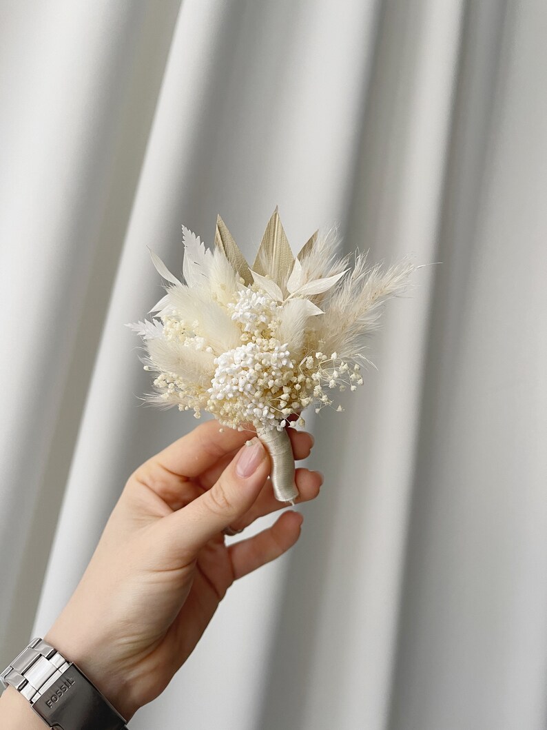 May include: A small bouquet of dried flowers in shades of white and beige, tied with a white ribbon. The bouquet includes pampas grass, baby's breath, and other dried foliage.