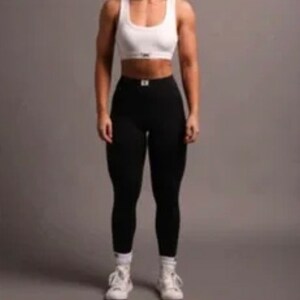 Op de afbeelding: Een witte sportbeha met een ronde hals en een klein rechthoekig label op de borst, gecombineerd met een zwarte legging met hoge taille met een bijpassend label op de tailleband, en witte sportschoenen met witte sokken.