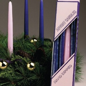 Puede incluir: Un juego de cuatro velas de Adviento de 30 cm en una caja rosa y morada. La caja dice "Velas de Adviento" y "Hecha a mano". Las velas están dispuestas en una corona con ramas de pino verdes y adornos dorados.