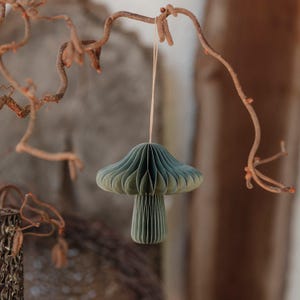 Könnte beinhalten: Ein dekorativer Papierpilz in Grüntönen hängt an einem Faden, der an einem Ast befestigt ist. Der Pilz hat ein geschichtetes, plissiertes Design, und der Stiel ist einfarbig grün. Der Hintergrund ist verschwommen, mit Andeutungen von Braun und Beige.