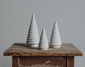 May include: Three white ceramic Christmas trees of varying sizes on a rustic wooden table. The trees are all in the shape of a traditional Christmas tree with a pointed top and layered branches.