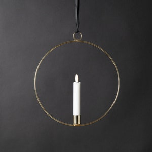 Könnte beinhalten: Ein goldfarbener Kerzenhalter aus Metall mit einem runden Design. Der Halter ist an einem schwarzen Band befestigt und hat eine weiße Kerze.