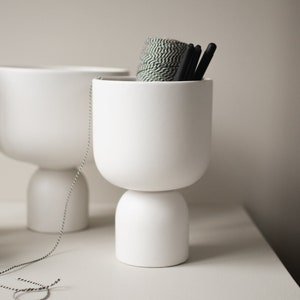 Könnte beinhalten: Zwei weiße Keramik-Blumentöpfe mit abgerundeter Basis und einem breiten, zylindrischen Oberteil. Der Topf rechts enthält eine Rolle grünes und weißes Garn und schwarze Stifte.