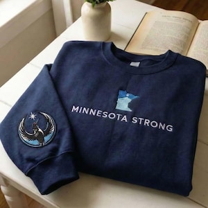 Peut inclure: Sweat-shirt bleu marine avec l'inscription blanche brod&eacute;e "MINNESOTA STRONG". Un &eacute;cusson avec un motif d'oiseau est pr&eacute;sent sur la manche gauche. Le sweat-shirt pr&eacute;sente &eacute;galement un contour de l'&Eacute;tat du Minnesota.