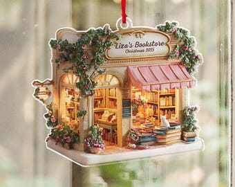 May include: A detailed ornament of a bookstore, with a pink awning, floral accents, and the text "Liza's Bookstore Christmas 2025." The miniature shop is filled with books and has a small white cat sitting on a stack of books.