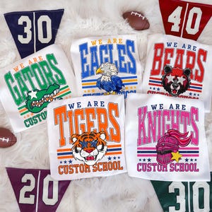 May include: White t-shirts with team mascots and text, including a gator, eagle, bear, tiger, and knight. Each shirt has the words "We Are" above the mascot and "Custom School" below. Pennants with numbers 20, 30, and 40 are also visible.