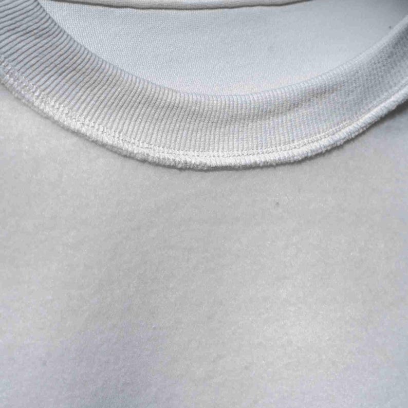 Peut inclure: Gros plan sur un sweat-shirt gris clair. Le col ras du cou c&ocirc;tel&eacute; est d'une nuance de gris l&eacute;g&egrave;rement plus fonc&eacute;e et est cousu sur le corps du v&ecirc;tement. Le tissu a une texture douce et l&eacute;g&egrave;rement floue.