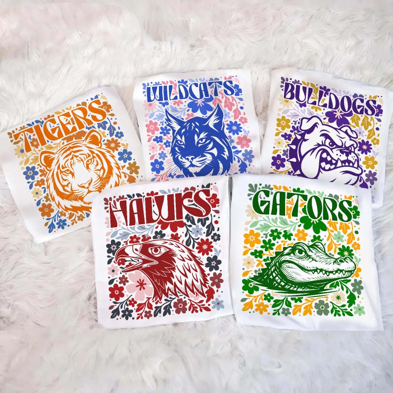 May include: Five white t-shirts with school mascot designs. The shirts feature a tiger, wildcat, bulldog, hawk, and alligator, each surrounded by floral patterns. The text on the shirts reads "Tigers", "Wildcats", "Bulldogs", "Hawks", and "Gators".
