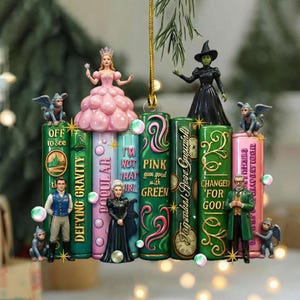 Può includere: Un ornamento decorativo con personaggi e citazioni del musical "Wicked" e del libro "Il Mago di Oz". L'ornamento include figure di Glinda, Elphaba e altri personaggi sopra strutture colorate a forma di libro con testo.