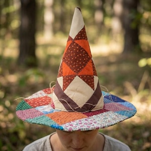 Puede incluir: Un sombrero cónico construido con parches de tela acolchada. La copa alta presenta un patrón geométrico de estrellas en tonos naranja, marrón y crema. El ala ancha y circular está compuesta por varios cuadrados de tela floral y estampada.