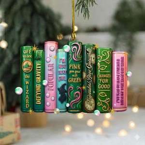 May include: A colourful book-shaped ornament with titles like "Defying Gravity" and "Popular." The ornament is suspended by a gold string and decorated with sparkling accents. The books are in various colours, including green, pink, and teal.