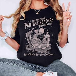 Pu&ograve; includere: Maglietta nera con una grafica di un libro, un drago e una candela. Il testo sulla maglietta recita "The Tortured Fantasy Readers Department" e "All's Fair In Love And Epic Tales".