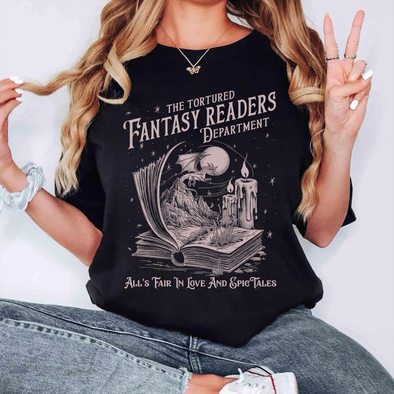 Pu&ograve; includere: Maglietta nera con una grafica di un libro, un drago e una candela. Il testo sulla maglietta recita "The Tortured Fantasy Readers Department" e "All's Fair In Love And Epic Tales".
