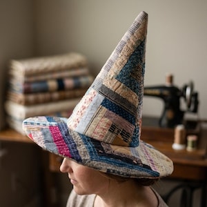 Puede incluir: Un sombrero alto y puntiagudo construido con varias tiras de tela en un diseño de patchwork. El sombrero presenta un ala ancha y circular y una corona cónica, mostrando una mezcla de patrones en azul, beige, crema y rojo apagado.