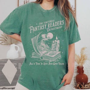 Pu&ograve; includere: Una maglietta verde con una stampa grafica bianca di un libro, un drago e candele. Il testo sulla maglietta recita "The Tortured Fantasy Readers Department" e "All's Fair in Love and Epic Tales".