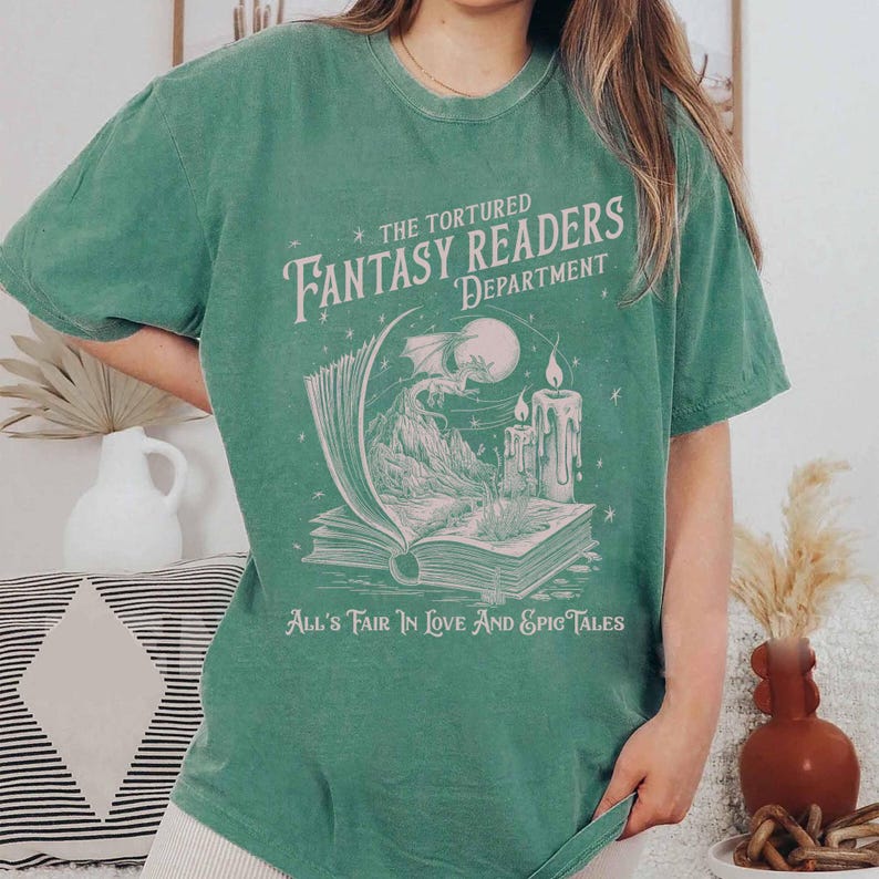 Pu&ograve; includere: Una maglietta verde con una stampa grafica bianca di un libro, un drago e candele. Il testo sulla maglietta recita "The Tortured Fantasy Readers Department" e "All's Fair in Love and Epic Tales".