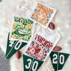 May include: White t-shirts with colorful floral designs and team mascots: a tiger, an alligator, and an eagle. Green felt pennants with yard line numbers and miniature footballs are also present.