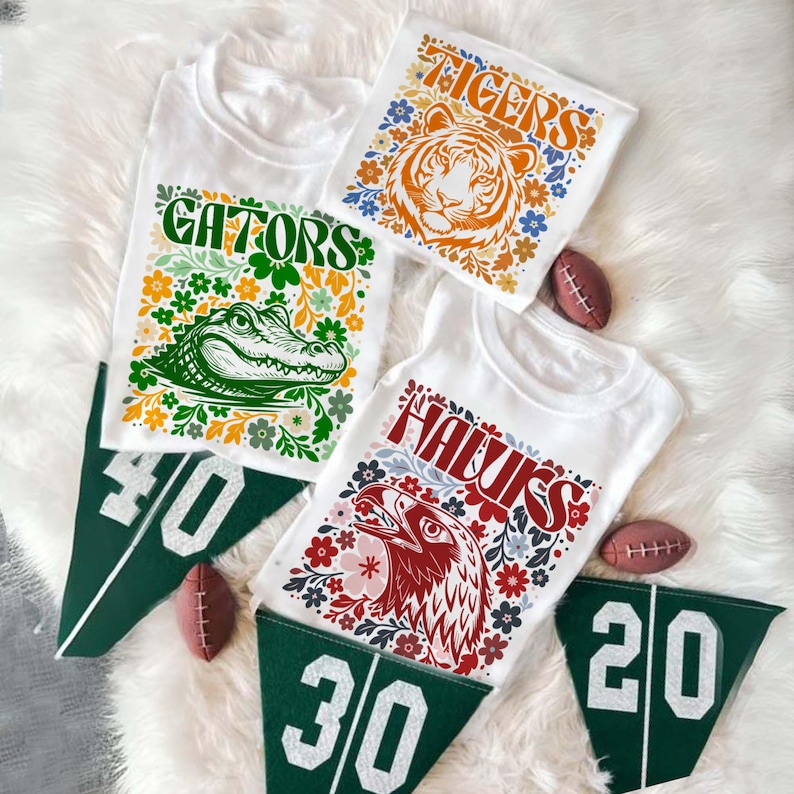 May include: White t-shirts with colorful floral designs and team mascots: a tiger, an alligator, and an eagle. Green felt pennants with yard line numbers and miniature footballs are also present.