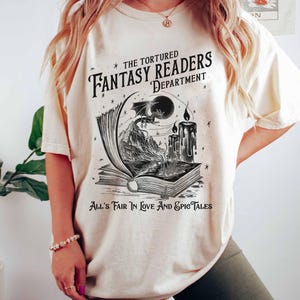 Pu&ograve; includere: Una maglietta bianca con un design grafico nero di un libro con un drago che vola fuori da esso. Il testo sulla maglietta recita "The Tortured Fantasy Readers Department" e "All's Fair In Love And Epic Tales".