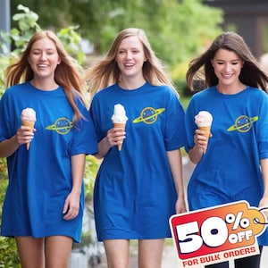Puede incluir: Tres personas con camisetas azules con un gráfico de planeta y sosteniendo conos de helado. Las camisetas tienen un anillo amarillo alrededor de un diseño de planeta. Un letrero en la esquina inferior derecha dice "50% DE DESCUENTO PARA PEDIDOS AL POR MAYOR".